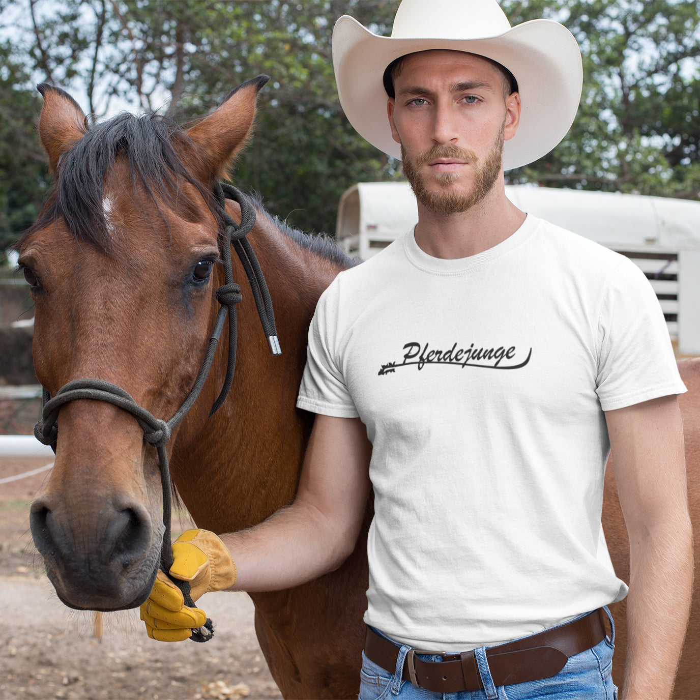 Ein Cowboy hält ein Pferd am Strick und trägt Cowboyhut und ein weißes Tshirt auf dem "Pferdejunge" steht