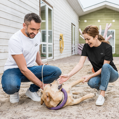 Ein junges Paar spielt mit einem Hund im Garten und trägt dabei 2 Varianten desselben Tshirts. Der Mann trägt ein weißes Tshirt auf dessen Brust ein kleiner schwarzer Cowboyhut zu sehen ist, die Frau trägt dasselbe Tshirt in schwarz