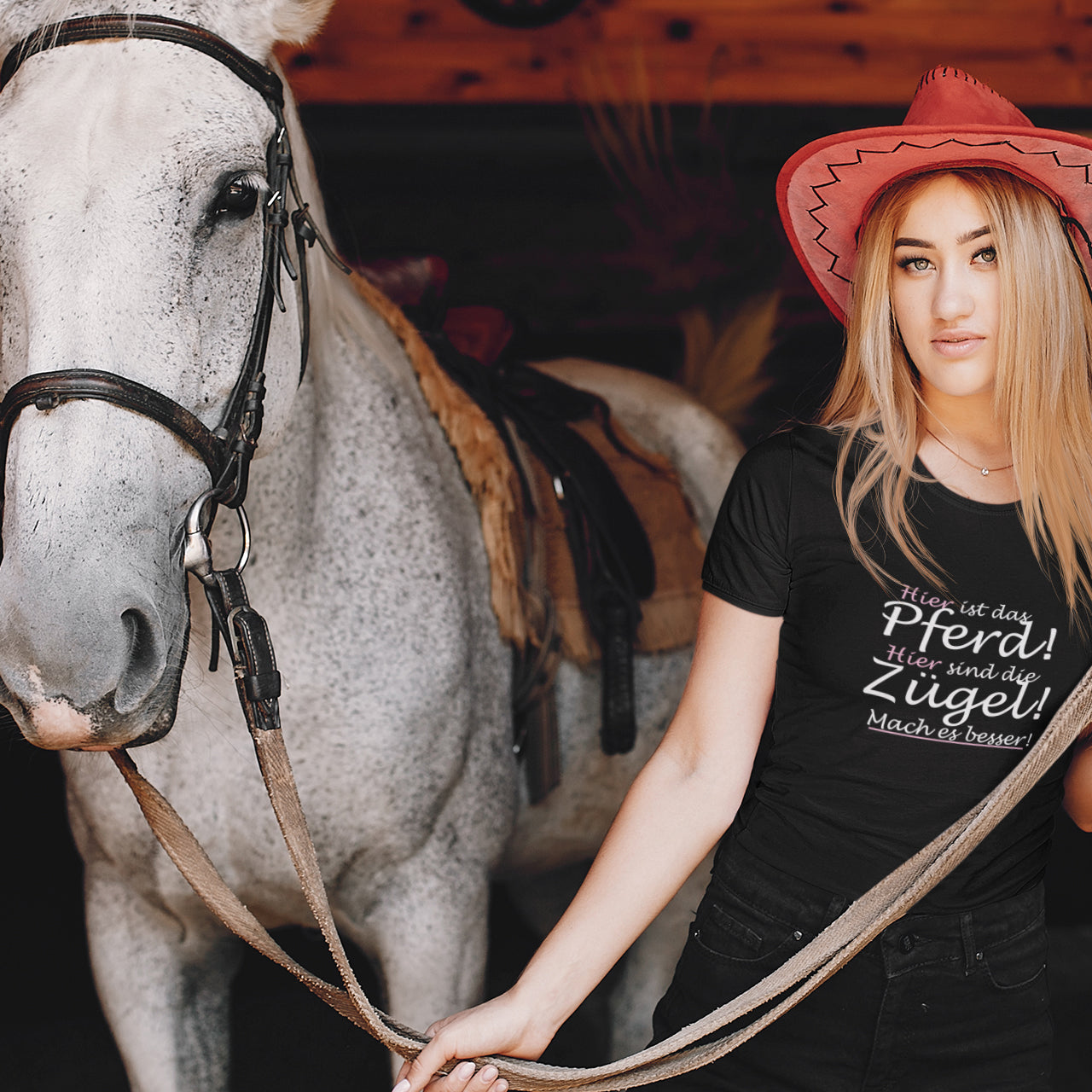 Eine junge Frau hält ein weißes Pferd am Halfter und trägt einen roten Cowboyhut sowie ein schwarzes Tshirt auf dem ein Spruch für Reiter abgebildet ist